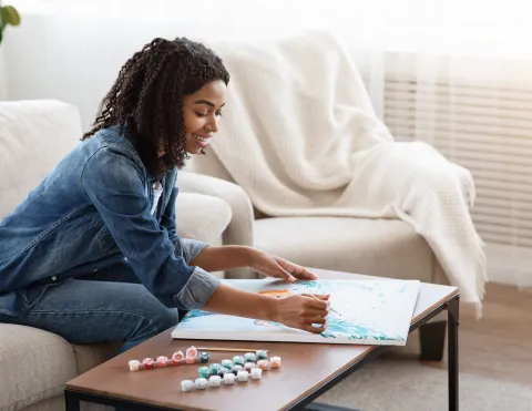 A Female using a paint brush on a canvas, with paint alongside her.