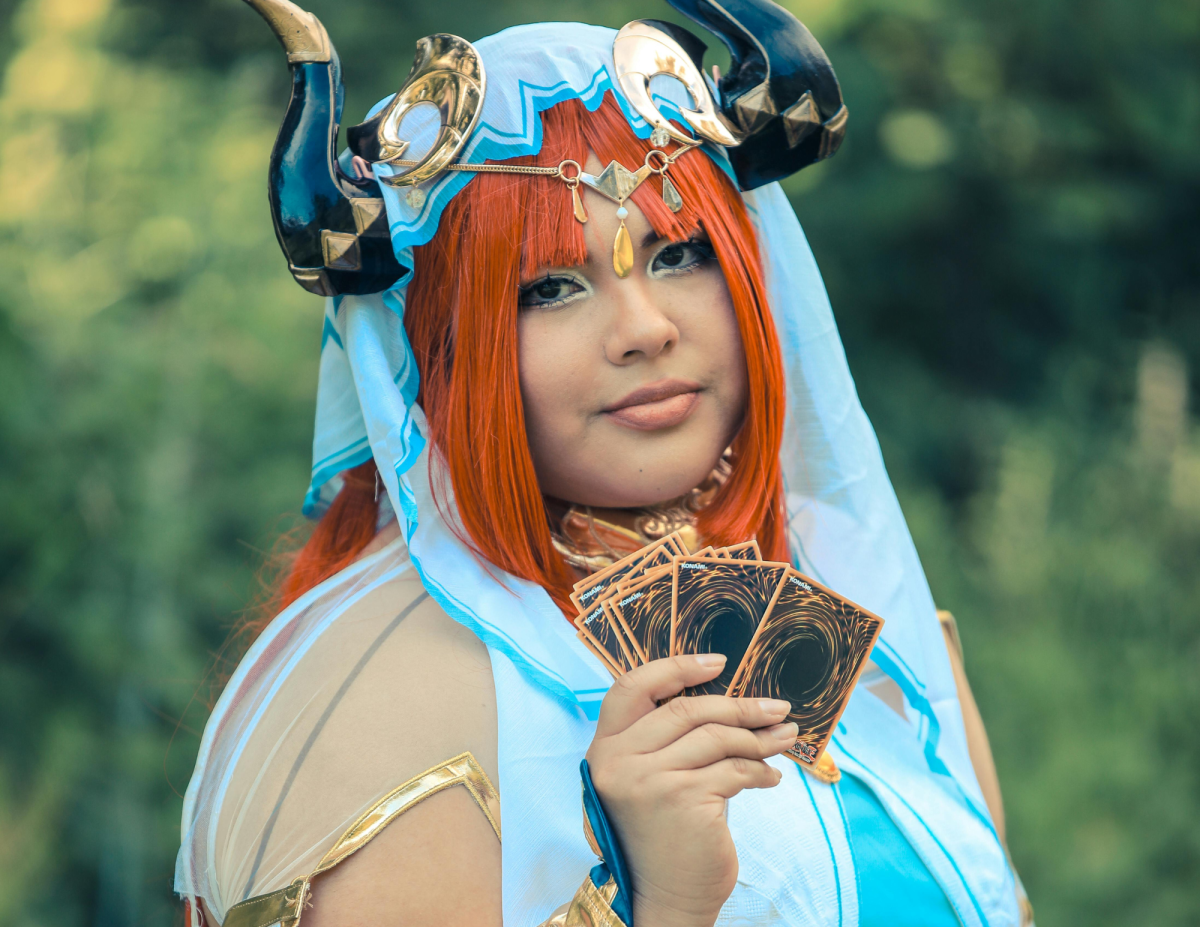 A Female cosplaying in a light blue cloak with horns, holding Yu-gi-oh cards. 